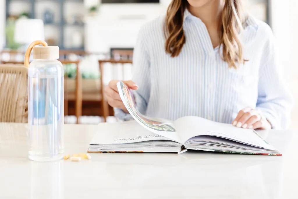 A woman in a striped shirt reading at a sunlit table, with a glass water bottle and a few supplement capsules beside her