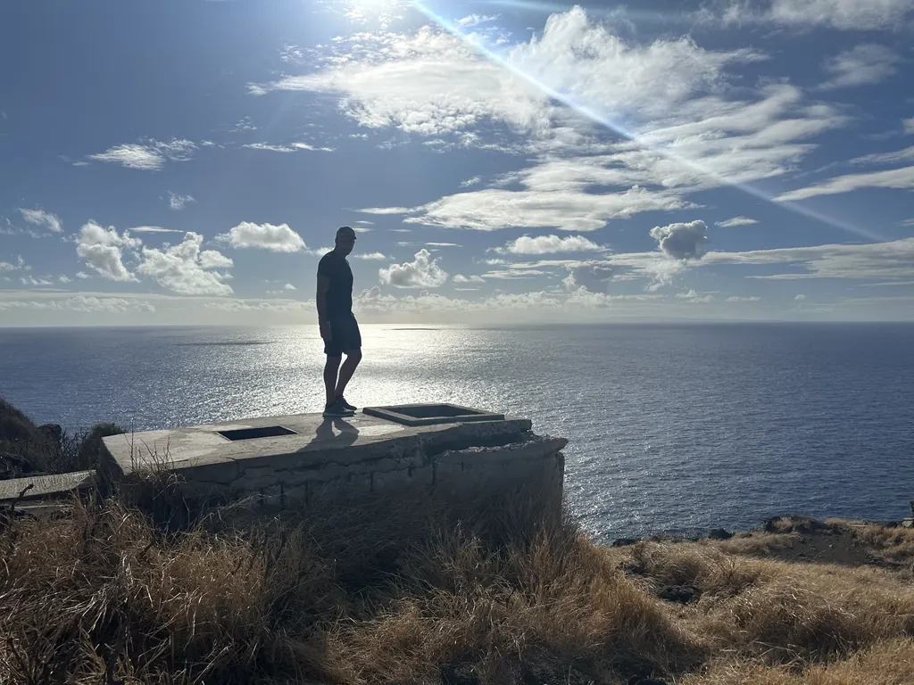 A person standing alone on a stone overlook, gazing out over a sunlit ocean and open sky