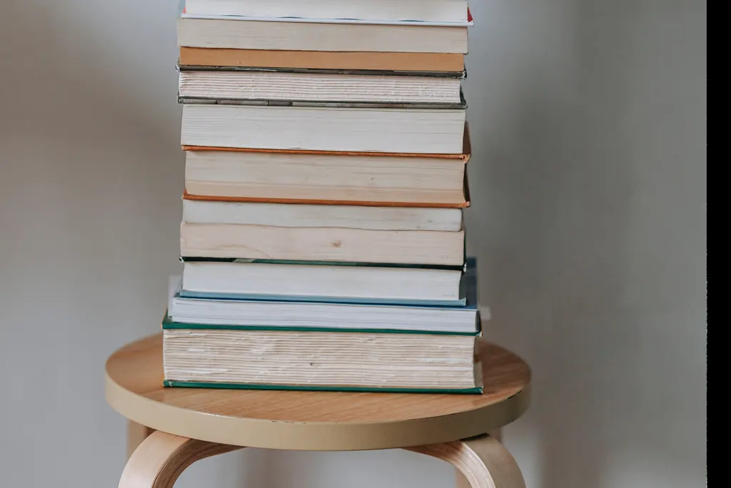 A tall stack of books resting on a wooden stool against a soft gray wall