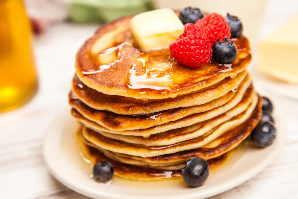 A stack of golden pancakes topped with butter, syrup, raspberries, and blueberries